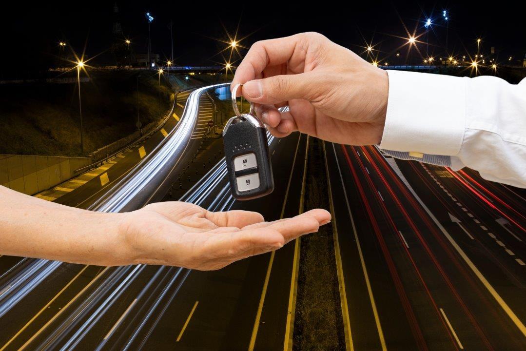 Hand giving car keys to another hand, with a blurred highway at night in the background, showing light trails from passing vehicles.