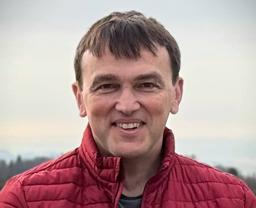 A person smiling outdoors, wearing a red quilted jacket, with a blurred natural background.